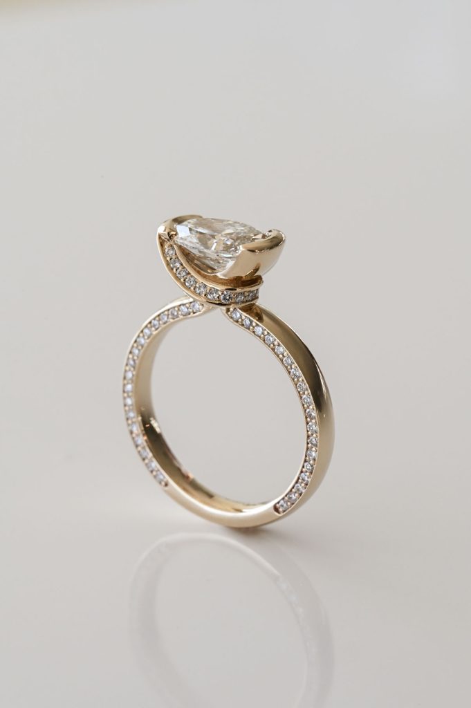 Yellow gold ring with a pear-shaped diamond set in an elevated semi-bezel and pavé diamond detailing along the band and under-gallery.